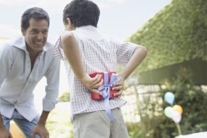 Man outdoors smiling with young boy hiding gift behind his back